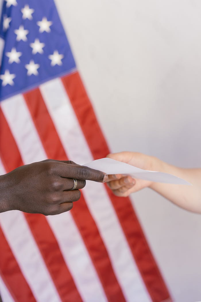 services-01 Two hands exchanging a paper in front of a blurred American flag background.